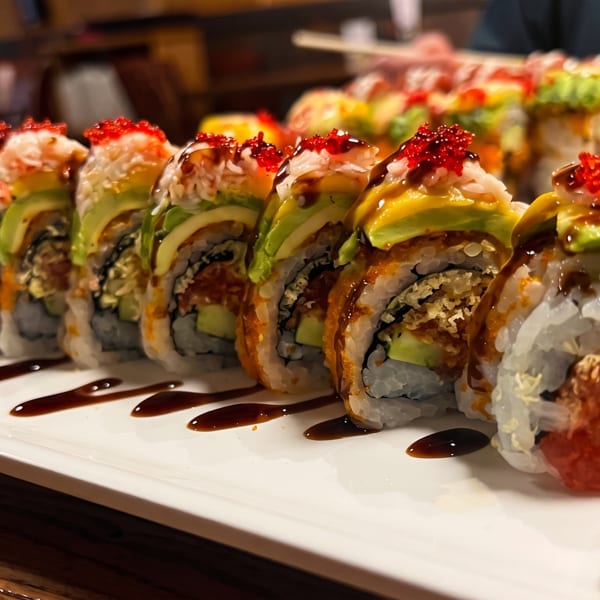 A long white plate of sushi rolls topped with avocado, red roe, and dark sauce.
